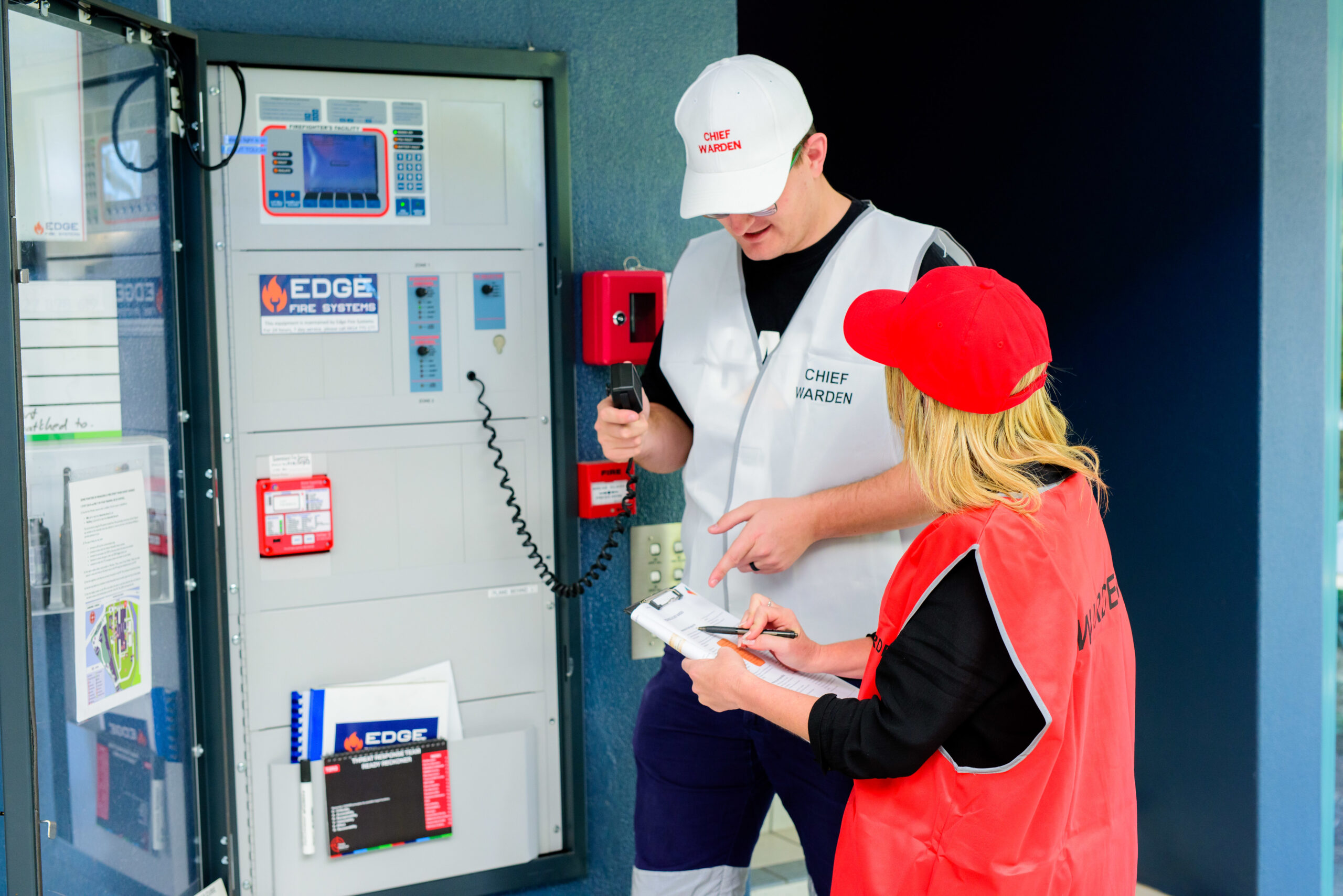 Chief Warden and an emergency officer reviewing fire safety procedures at a control panel during a training session, with the Chief Warden holding a radio and the officer taking notes on a clipboard, both wearing vests and caps that indicate their roles.