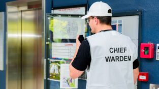 Chief Warden standing in front of a fire control panel, speaking into a radio during a training exercise, wearing a white vest and cap, with the fire alarm block plan visible in the background.